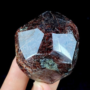 Könnte beinhalten: Ein facettierter, dunkelroter Kristall mit einer komplexen geometrischen Form. Der Kristall hat eine raue, strukturierte Oberfläche mit Bereichen in tiefem Rot und Braun. Zwei große, reflektierende Facetten sind sichtbar, die das Licht einfangen. Der Kristall wird vor einem schwarzen Hintergrund gehalten.
