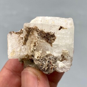 May include: A raw, natural crystal cluster. The mineral features a translucent, off-white color with areas of brown and tan matrix. The crystal has a rough, textured surface and a crystalline structure, showcasing its natural formation.