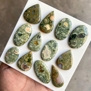 May include: A collection of polished gemstones in various shapes, including teardrop and oval. The stones feature a mix of green, brown, and cream colors with unique patterns. The gemstones are displayed on a white surface.