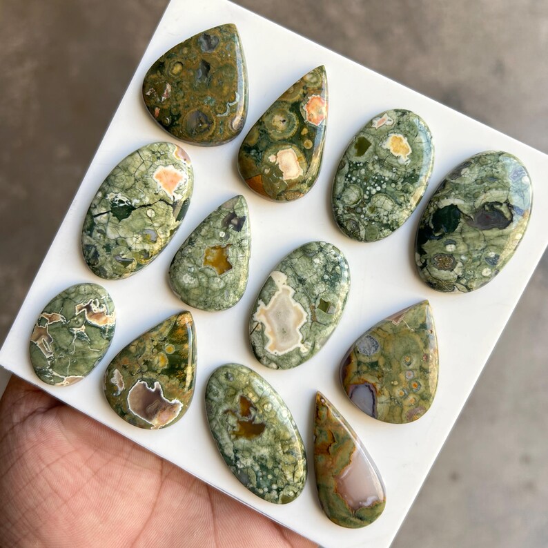 May include: A collection of polished gemstones in various shapes, including teardrop and oval. The stones feature a mix of green, brown, and cream colors with unique patterns. The gemstones are displayed on a white surface.