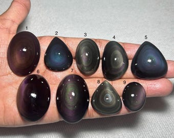 May include: Nine polished obsidian stones in various shapes, including oval, teardrop, and triangular. The stones display iridescent swirls of color, primarily purple, green, and black, with a glossy finish. The stones are displayed on a light-colored surface.