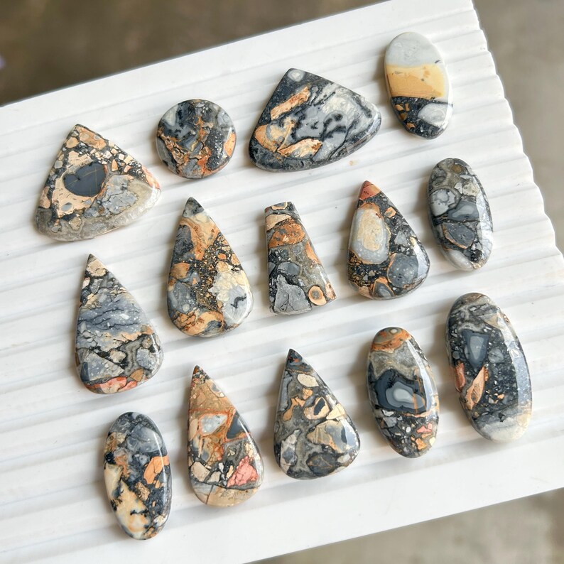 Puede incluir: Una colecci&oacute;n de piedras preciosas pulidas en varias formas, incluyendo gotas, &oacute;valos y tri&aacute;ngulos. Las piedras presentan una mezcla de colores gris, negro, naranja y crema, con patrones y texturas &uacute;nicos. Las piedras preciosas est&aacute;n dispuestas sobre una superficie blanca.