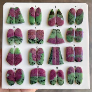 May include: A collection of sixteen pairs of ruby zoisite gemstones, each with a unique shape. The stones display a mix of deep red and green hues, with some featuring black inclusions. The gemstones are arranged on a white surface.