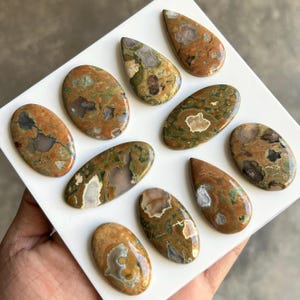 May include: A collection of polished gemstones in various shapes, including oval and teardrop. The stones feature a mix of earthy tones, with shades of brown, green, and cream. The gemstones are displayed on a white surface.