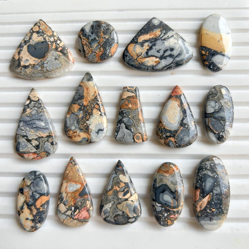 Puede incluir: Una colecci&oacute;n de quince piedras preciosas pulidas de diversas formas, incluyendo l&aacute;grimas, &oacute;valos y tri&aacute;ngulos. Las piedras presentan una mezcla de colores grises, negros, naranjas y crema con patrones intrincados. Las piedras preciosas son probablemente para la fabricaci&oacute;n de joyas o coleccionar.