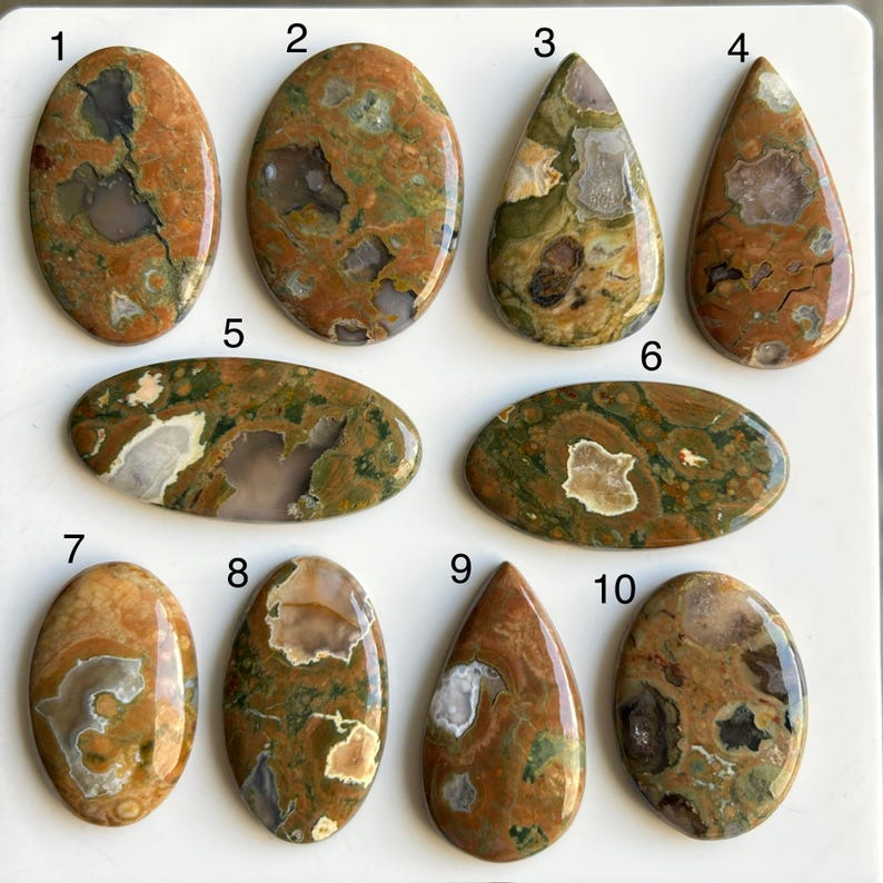 May include: A collection of ten polished gemstone cabochons in various shapes, including oval and teardrop. The stones feature a mix of earthy tones, with brown, green, and cream colors. Each stone has unique patterns and textures, suitable for jewelry making or collecting.