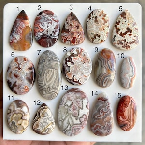 Puede incluir: Quince cabujones de piedras preciosas pulidas en varias formas, incluyendo ovaladas, en forma de lágrima y oblongas. Las piedras muestran una gama de colores, como marrón, rojo, blanco y gris, con patrones y texturas únicos. Las piedras preciosas están numeradas del 1 al 15.