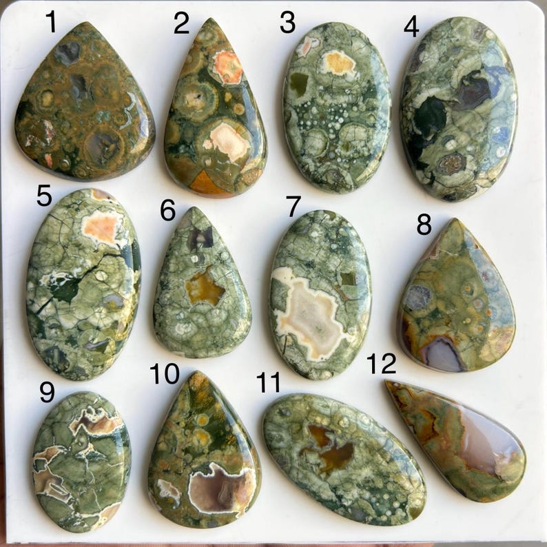 May include: A collection of twelve polished gemstones in various shapes, including teardrops and ovals. The stones display a range of colors, primarily greens, browns, and creams, with unique patterns and textures. Ideal for jewelry making or collecting.