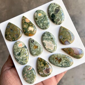 May include: A collection of polished gemstones in various shapes, including oval and teardrop. The stones feature a mix of green, brown, and cream colors with unique patterns. The gemstones are displayed on a white surface.