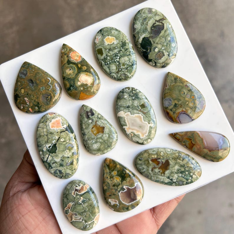 May include: A collection of polished gemstones in various shapes, including oval and teardrop. The stones feature a mix of green, brown, and cream colors with unique patterns. The gemstones are displayed on a white surface.
