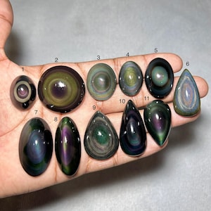 Puede incluir: Una colección de once piedras preciosas de obsidiana pulidas en varias formas, incluyendo ovaladas, redondas y en forma de lágrima. Las piedras muestran bandas iridiscentes de color, con tonos de verde, púrpura y dorado. Las piedras preciosas se sostienen en una mano.