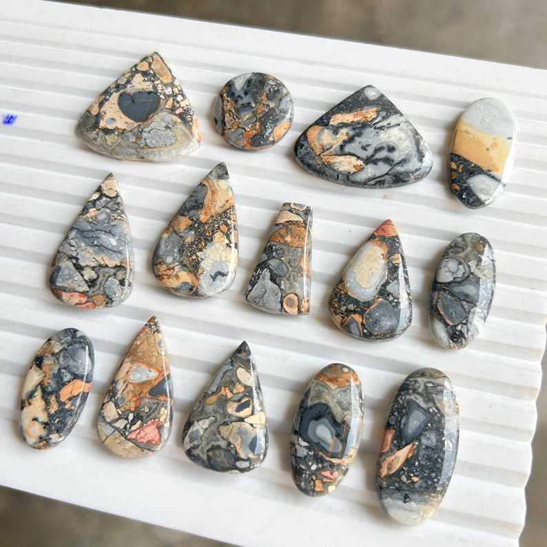Puede incluir: Una colecci&oacute;n de piedras preciosas pulidas en varias formas, incluyendo l&aacute;grimas, &oacute;valos y tri&aacute;ngulos. Las piedras presentan una mezcla de colores, como gris, beige, naranja y negro, con patrones y texturas &uacute;nicos. Las piedras preciosas se exhiben sobre una superficie blanca.