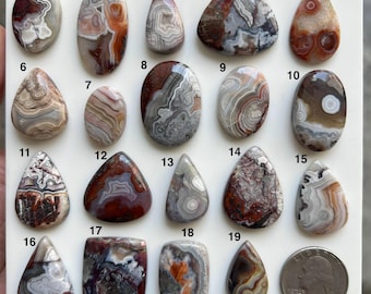 May include: A collection of nineteen polished, multi-colored agate cabochons arranged on a white display board, numbered one through nineteen, featuring varied swirling patterns in shades of brown, gray, white, and red, alongside a U.S. quarter coin.