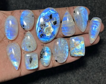 May include: A collection of twelve polished moonstone cabochons in various shapes, including oval, pear, and rectangular cuts. Each stone displays a translucent white base with iridescent blue flashes and dark mineral inclusions, numbered 1 through 12.