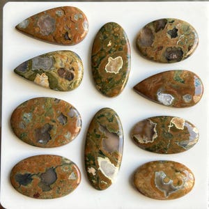 May include: A collection of polished gemstones in various shapes, including oval and teardrop. The stones display a mix of earthy tones, with brown, green, and cream colors. Some stones have visible patterns and inclusions, showcasing natural variations.