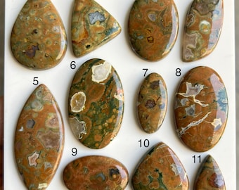 May include: A collection of eleven polished gemstones in various shapes, including teardrop, triangular, and oval. The stones display a mix of earthy tones, with brown, green, and beige patterns. The gemstones are likely for jewelry making or collecting.