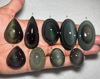 May include: Nine polished obsidian gemstones in various shapes, including teardrop and oval. The stones display swirling patterns of dark green, brown, and black, with a glossy finish. The gemstones are held in a hand.