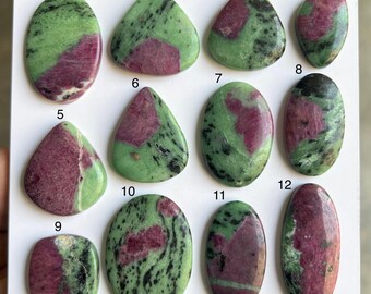 Può includere: Assortimento di dodici pietre preziose di zoisite lucidata con inclusioni di rubino. Le pietre variano in forma, tra cui ovale, a goccia e rettangoli arrotondati. I colori sono un mix di verde e rosso intenso.