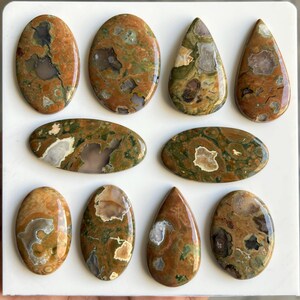 May include: A collection of polished gemstones in various shapes, including oval and teardrop. The stones feature a mix of earthy tones, such as brown, green, and beige, with unique patterns and inclusions. The gemstones are displayed on a white surface.