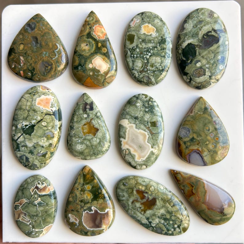 May include: A collection of twelve polished gemstones in various shapes, including teardrop and oval. The stones feature a mix of green, brown, and cream colors with unique patterns and textures. These natural stones are ideal for jewelry making or collecting.