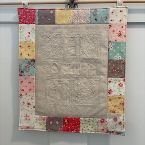 以下が含まれることがあります： 裁縫をテーマにした画像と「QUILT」の文字が刺繍された、中央のリネンパネルを備えたキルト壁掛け。 パネルは、ピンク、黄色、青など、さまざまな色の花柄と無地の生地の正方形のパッチワークボーダーで縁取られています。