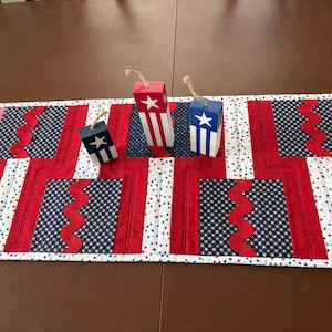 Puede incluir: Un camino de mesa de tela rectangular con un diseño de patchwork en patrones rojos, blancos y azules con motivos de estrellas y puntos, acentuado por un borde ondulado rojo. Tres pequeños bloques de madera rectangulares con lazos de yute.