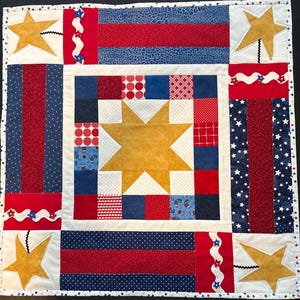 May include: A patriotic quilt featuring a central gold star surrounded by red, white, and blue patchwork squares. The design includes gold stars, stripes, and wavy accents, all framed by a white border with star-shaped embellishments. The quilt is a square shape.