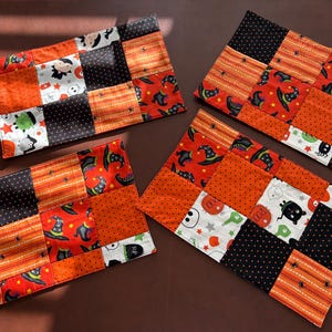 Puede incluir: Tres manteles individuales rectangulares de tela con un diseño de patchwork de cuadrados de algodón en naranja, negro y blanco. Los patrones incluyen ilustraciones de sombreros de bruja, fantasmas, arañas y calabazas. Cada mantel está acolchado.