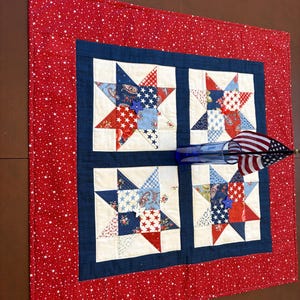 May include: A square fabric quilt featuring four star-shaped blocks in red, white, and blue patterns, surrounded by a dark blue border and a red outer border with white stars. A blue glass vase holds a small American flag on the quilt surface.