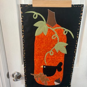 May include: A fabric wall hanging with a large orange pumpkin design, green and white striped leaves, and a brown stem. A black bird, an acorn, and a leaf are appliqued on a black background. The edges have a patterned border, perfect for autumn decor.