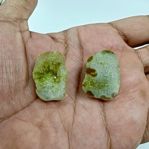 Könnte beinhalten: Zwei rohe, ungeschnittene Achatscheiben. Eine ist hellgrün mit einem kristallinen Zentrum, die andere eine Mischung aus Weiß, Grün und Braun. Diese Natursteine eignen sich ideal für die Schmuckherstellung oder zum Sammeln. Jede Scheibe hat eine einzigartige Form.