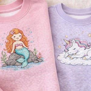 May include: Two sweatshirts, one pink and one lavender, featuring embroidered designs. The pink sweatshirt has a mermaid design, and the lavender one has a sleeping unicorn on a cloud. Both have a crew neck.