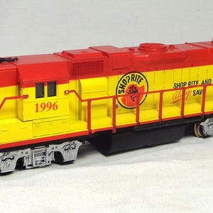 May include: A model diesel locomotive train engine painted in bright yellow and red. The side features the ShopRite logo, the text 1996, and a 50th Anniversary emblem. It has silver-coloured wheels and undercarriage details with red railings.