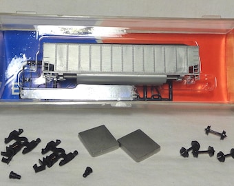 May include: A model train car in a clear plastic case with blue and orange accents. The silver car has a rectangular shape with detailed paneling. Several small black train parts and two gray rectangular pieces are also included.