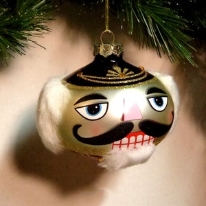 Puede incluir: Un adorno de Navidad de vidrio con forma de cabeza de cascanueces. El adorno es plateado con detalles negros, incluyendo un bigote y cejas. El cascanueces tiene una nariz rosa y un borde de piel blanca.