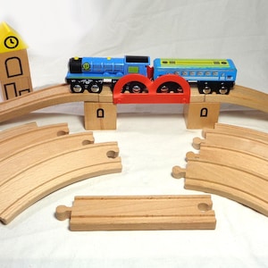 May include: A wooden toy train set featuring a blue and green locomotive with a matching passenger car, a red bridge, curved and straight wooden tracks, two green tree figures, a clock tower block, and a stacked block tower structure.