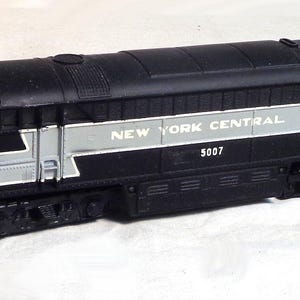 May include: A black and silver model train locomotive with the words "NEW YORK CENTRAL" and the number "5007" printed on the side. The model train is a vintage collectible.