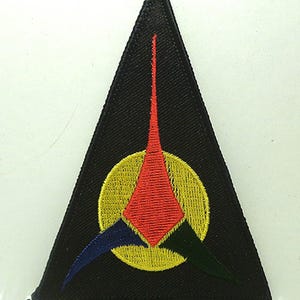 May include: A black triangular patch with an embroidered yellow circle and a red, green, and blue triangular design inside.