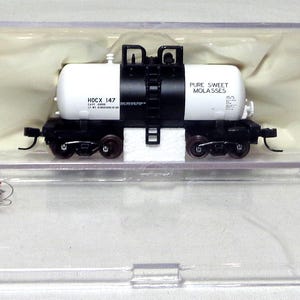 May include: A model train tank car in a clear plastic case. The tank car is white with black accents and the words "PURE SWEET MOLASSES" and "HOCK 147" printed on it. The model is on a black chassis with wheels.