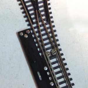 Op de afbeelding: Een close-up van een modelspoorwegschakelaar met een zwarte plastic bedieningshendel. Het spoor is gemaakt van zwart plastic met gouden metalen rails. De schakelaar is ontworpen om treinen van spoor te laten wisselen.