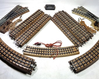 Puede incluir: Un conjunto de piezas de vías de tren a escala que incluye dos desvíos, varias secciones rectas y curvas con traviesas de madera y rieles metálicos, una pequeña caja de control negra y cableado eléctrico con conectores rojos y marrones.