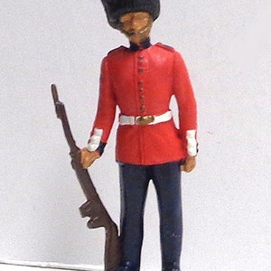 May include: A toy soldier figurine wearing a red uniform with a black hat and holding a rifle. The soldier is standing on a gray base.