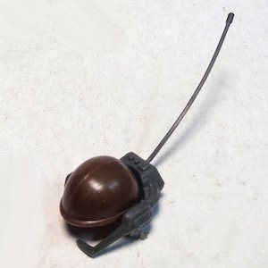 May include: A brown, helmet-shaped toy with a dark gray antenna and attached components. The toy is likely a miniature military or action figure accessory, designed to be held or worn by a small figure. The antenna extends upwards.