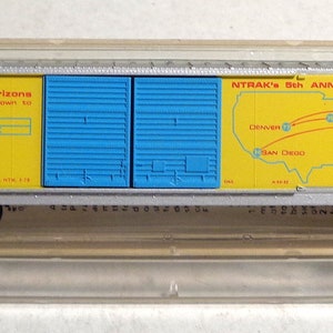 May include: A yellow and blue model train car with the text "NTRAK's 5th Anniversary" and a map of the United States showing the route of the train. The car also has the text "broaden your horizons scale down to NTRAK" and "NTK 1978".