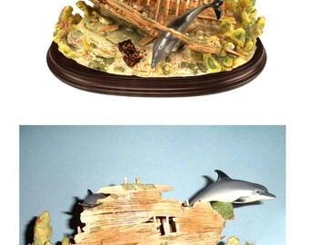 May include: A figurine of a shipwreck with three dolphins swimming around it. The shipwreck is made of wood and has a weathered look. The dolphins are painted in shades of gray and white. The figurine is set on a brown base.