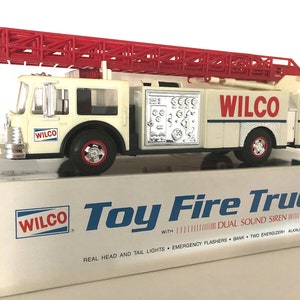 May include: A white toy fire engine with a red ladder, the words "WILCO" on the side, and a box with the words "WILCO Toy Fire Truck" on it. The box also says "WITH DUAL SOUND SIREN, REAL HEAD AND TAIL LIGHTS, EMERGENCY FLASHERS, BANK TWO ENERGIZER ALKALINE BATTERIES."
