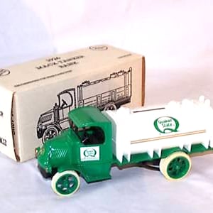 Op de afbeelding: Een groene 1926 Mack Tanker Truck spaarpot met een witte tank en gouden versieringen. De tank heeft een groen "Quaker State" logo met een grote "Q". De vrachtwagen staat op een wit oppervlak.