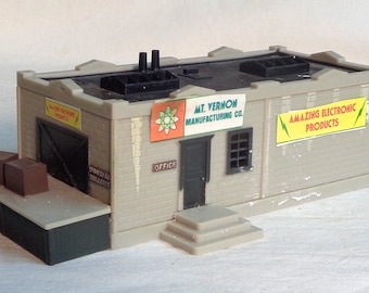 May include: A gray toy building with a black roof and a sign that reads "Mt. Vernon Manufacturing Co.". There are two boxes on a loading dock in front of the building.