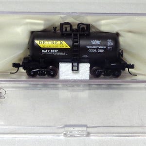 May include: A black model train tank car with the text "DETREX" and "GATX 9537" on a yellow label. The model is in a clear plastic case with the "ATLAS" logo and an "age 14+" sticker.