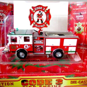 Peut inclure: Un camion de pompiers rouge et blanc avec une cabine verte, une échelle argentée et un '99' sur le côté. Le camion de pompiers est dans une vitrine en plastique transparent avec un fond rouge et blanc sur le thème de Noël. La boîte porte les mots 'CODE 3' et 'DIE-CAST COLLECTIBLES'.